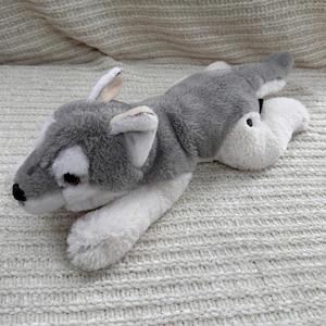 May include: A plush toy wolf in a lying position, featuring a gray and white color scheme. The wolf has a black nose, brown eyes, and white accents on its ears and paws. The toy is made of soft, fluffy material.