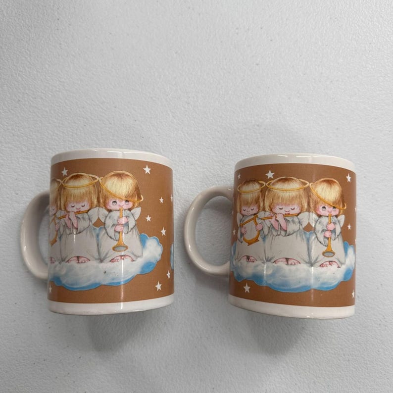 Puede incluir: Dos tazas de cer&aacute;mica con un fondo marr&oacute;n que presentan ilustraciones de &aacute;ngeles querubines tocando la trompeta y la flauta. Los &aacute;ngeles est&aacute;n representados en una nube con estrellas. Las tazas tienen asas y bordes blancos.