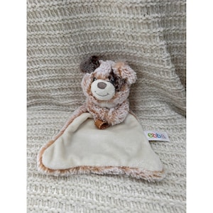 以下が含まれることがあります： 茶色とクリーム色の犬の形をした、柔らかいぬいぐるみベビーコンフォーター。 犬は、笑顔の表情と小さな茶色の革のタグが付いたステッチされた顔をしています。 コンフォーターは柔らかく、豪華な質感で、クリーム色の毛布が付いています。