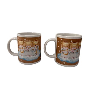 Puede incluir: Dos tazas de cer&aacute;mica blanca con un fondo marr&oacute;n que presenta un dise&ntilde;o de &aacute;ngeles querubines tocando instrumentos musicales. Los &aacute;ngeles visten t&uacute;nicas blancas y tienen halos dorados. Estrellas blancas est&aacute;n dispersas por el fondo marr&oacute;n.