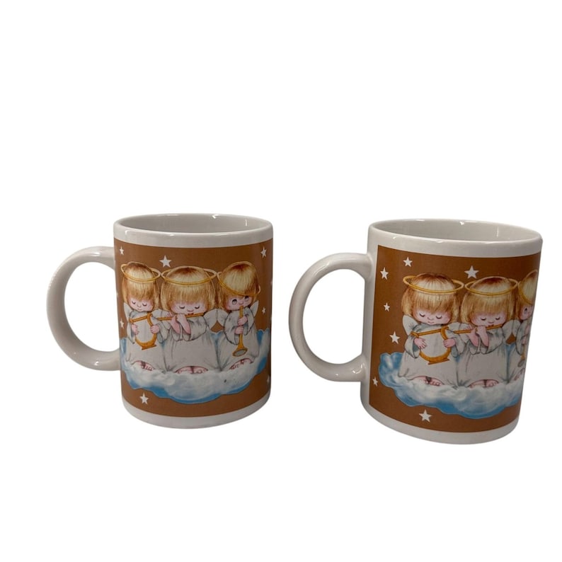 Puede incluir: Dos tazas de cer&aacute;mica blanca con un fondo marr&oacute;n que presenta un dise&ntilde;o de &aacute;ngeles querubines tocando instrumentos musicales. Los &aacute;ngeles visten t&uacute;nicas blancas y tienen halos dorados. Estrellas blancas est&aacute;n dispersas por el fondo marr&oacute;n.