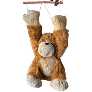 May include: A plush monkey toy with brown and tan fur, featuring long arms suspended by white strings from a horizontal wooden bar. The monkey has a light tan face, dark eyes, a black nose, and large hands and feet with stitched detailing.