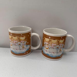 Puede incluir: Dos tazas de cer&aacute;mica con una banda marr&oacute;n con ilustraciones de &aacute;ngeles querubines tocando instrumentos sobre nubes. Las tazas tienen interiores y asas blancas, con peque&ntilde;as estrellas blancas.