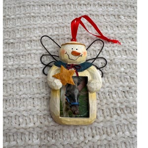 May include: A snowman-shaped Christmas ornament with a photo frame. The ornament is painted in cream, with a carrot nose, black eyes, and a red ribbon. It has wire wings and a gold star. The frame holds a photo of a horse.