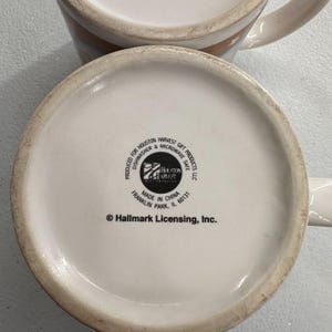 Puede incluir: Dos tazas de cer&aacute;mica blanca con asa. La base de las tazas muestra el texto "PRODUCED FOR HOUSTON HARVEST DISHWASHER & MICROWAVE SAFE LLC" y "MADE IN CHINA". Las tazas son de Hallmark Licensing, Inc.