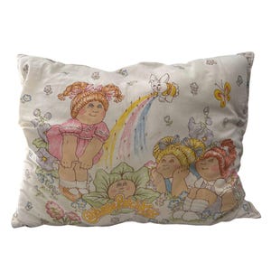Puede incluir: Una funda de almohada blanca con una colorida ilustración de Cabbage Patch Kids. El diseño incluye varias muñecas, un arcoíris, un conejito y flores. El texto "Cabbage Patch Kids" también es visible.