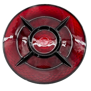May include: A round, ruby red serving dish with a divided design. The dish has a central circular compartment and four radiating sections, all in black. The red sections have a textured, diamond pattern.