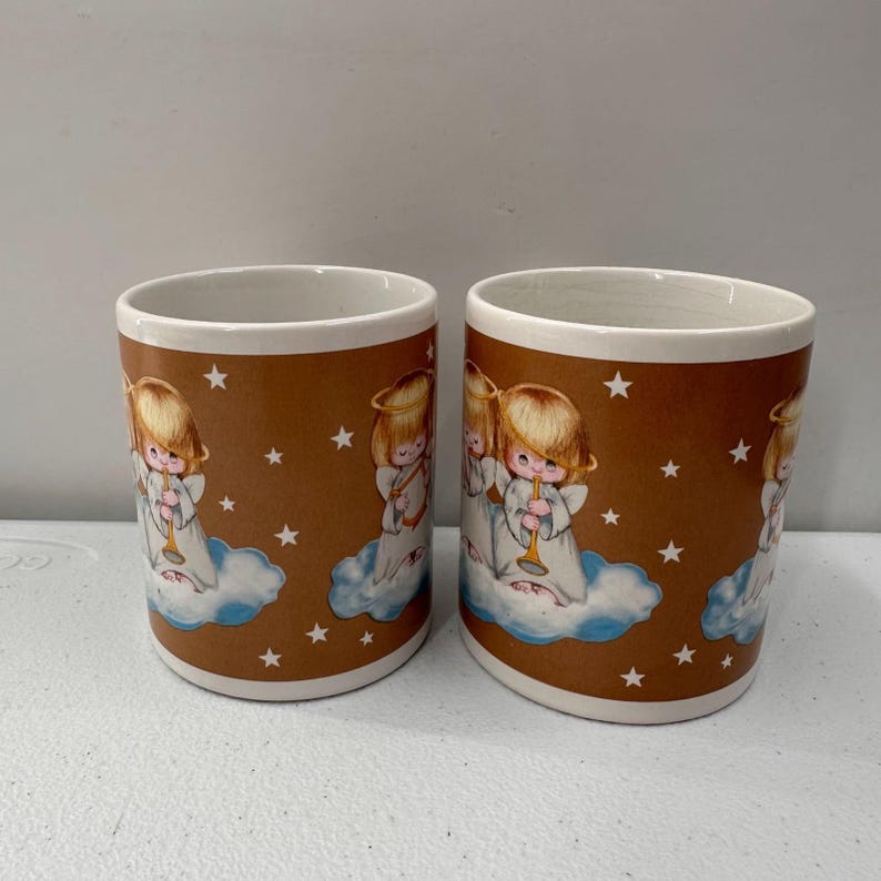 Puede incluir: Dos tazas de cer&aacute;mica con fondo marr&oacute;n e interior blanco. Cada taza presenta una ilustraci&oacute;n repetida de un &aacute;ngel querub&iacute;n tocando la trompeta sobre una nube, con estrellas blancas dispersas. Las tazas tienen una est&eacute;tica vintage.