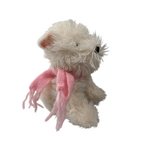 May include: A plush, white stuffed animal dog with a pink scarf. The dog has black eyes and a fluffy, textured coat. The pink scarf is knitted and has fringed ends. The toy is set against a white background.
