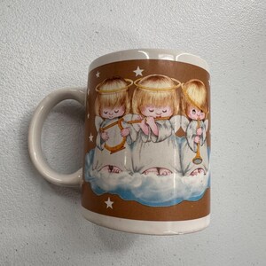 Puede incluir: Taza de cer&aacute;mica blanca con una banda marr&oacute;n que presenta tres figuras de querubines tocando instrumentos musicales. Los &aacute;ngeles est&aacute;n representados sobre una nube, con estrellas alrededor. La taza tiene asa y borde blancos.