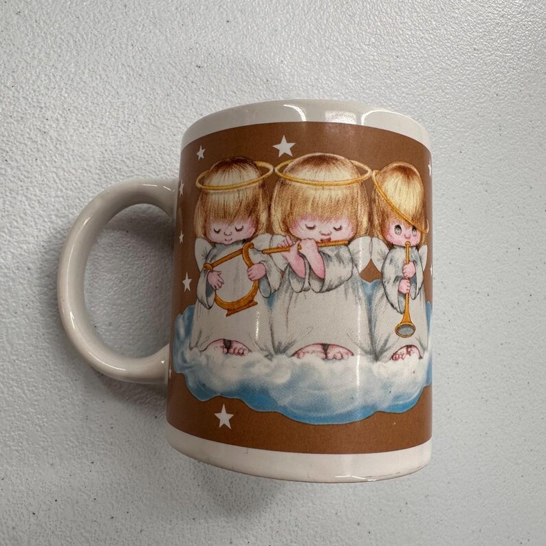 Puede incluir: Taza de cer&aacute;mica blanca con una banda marr&oacute;n que presenta tres figuras de querubines tocando instrumentos musicales. Los &aacute;ngeles est&aacute;n representados sobre una nube, con estrellas alrededor. La taza tiene asa y borde blancos.