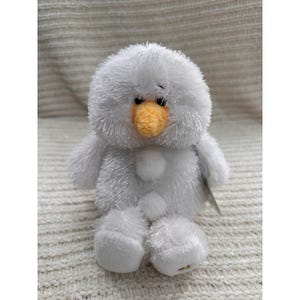 May include: A plush snowman toy with a fluffy white body, arms, and legs. It has a round head, a yellow beak, and two small black eyes. The snowman has three white buttons down its front and is sitting on a white, textured surface.