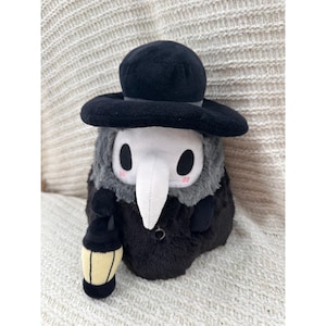 Puede incluir: Un peluche que representa a un médico de la peste, con sombrero negro, máscara de pico y abrigo largo. El juguete tiene una combinación de colores gris y negro y sostiene una linterna. El peluche tiene mejillas rosadas y una expresión linda.