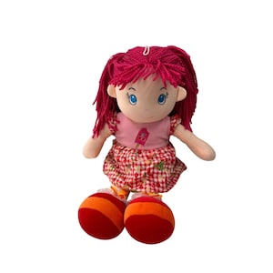 Pode incluir: Boneca de pano feita à mão com cabelo de fio vermelho e olhos azuis. A boneca usa um top rosa com um desenho de picolé e uma saia xadrez vermelha e branca. Os pés da boneca são vermelhos com listras laranja.