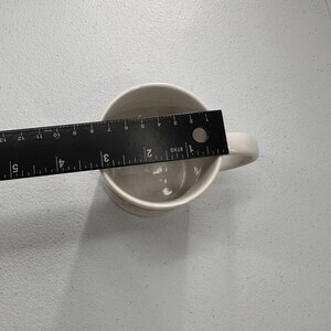 Puede incluir: Una taza de cer&aacute;mica color crema con asa. Una regla negra se coloca dentro de la taza, midiendo aproximadamente 7.6 cm de ancho. La taza est&aacute; sobre una superficie gris claro.