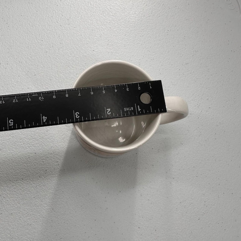 Puede incluir: Una taza de cer&aacute;mica color crema con asa. Una regla negra se coloca dentro de la taza, midiendo aproximadamente 7.6 cm de ancho. La taza est&aacute; sobre una superficie gris claro.