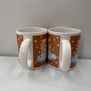 Puede incluir: Dos tazas de cer&aacute;mica blanca con un fondo marr&oacute;n que presenta un dise&ntilde;o de &aacute;ngel sobre una nube, con halos dorados y estrellas blancas. Cada taza tiene un asa blanca.