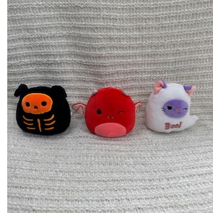 May include: Three plush toys in the shape of Halloween characters. A black skeleton, a red devil with wings, and a white ghost cat with the word "Boo!" on it. The toys are on a textured, light grey surface.