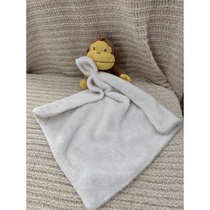 May include: A white plush baby security blanket with a small brown and yellow monkey toy attached. The blanket is square-shaped and has a soft, fuzzy texture. The monkey has a stitched face.