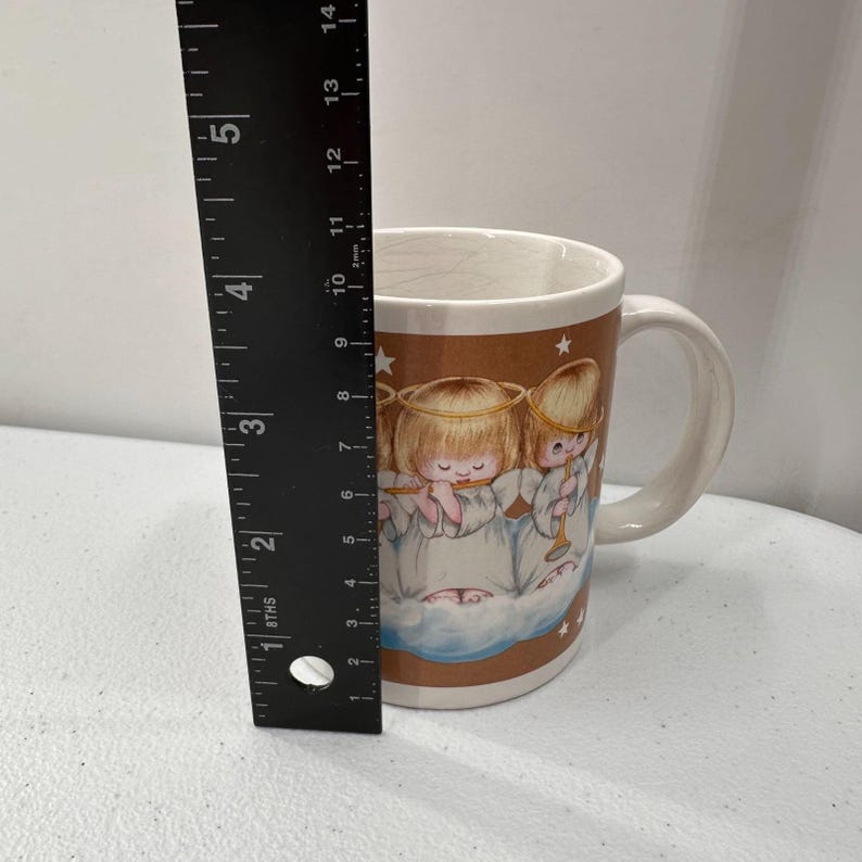 Puede incluir: Una taza de cer&aacute;mica con una banda marr&oacute;n con ilustraciones de &aacute;ngeles querubines tocando instrumentos musicales. La taza tiene un interior y un asa blancos. La taza mide aproximadamente 10 cm de alto.