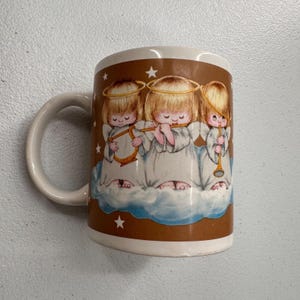 Puede incluir: Taza de cer&aacute;mica blanca con una banda marr&oacute;n que presenta a tres querubines tocando instrumentos musicales. Los querubines tienen cabello rubio, halos y t&uacute;nicas blancas, de pie sobre una nube. La taza tiene un asa blanca y un interior blanco.