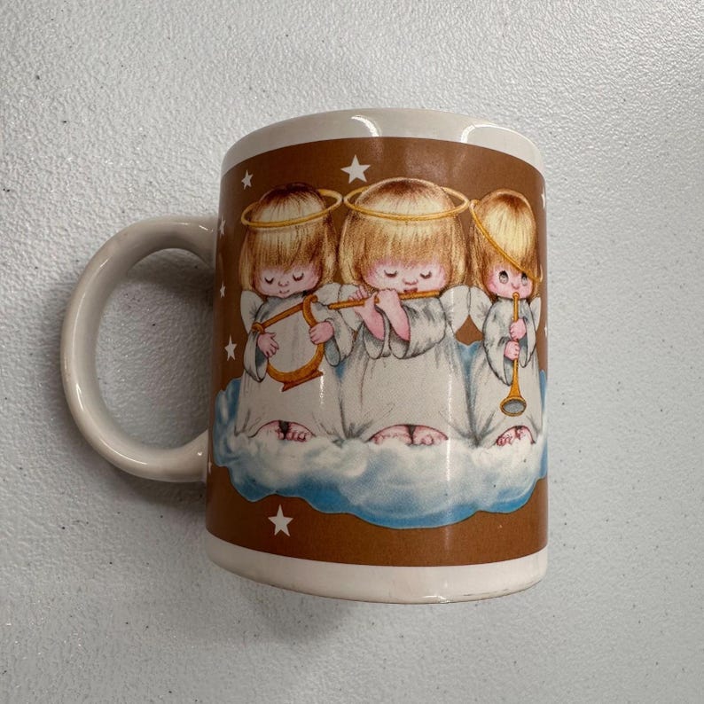 Puede incluir: Taza de cer&aacute;mica blanca con una banda marr&oacute;n que presenta a tres querubines tocando instrumentos musicales. Los querubines tienen cabello rubio, halos y t&uacute;nicas blancas, de pie sobre una nube. La taza tiene un asa blanca y un interior blanco.