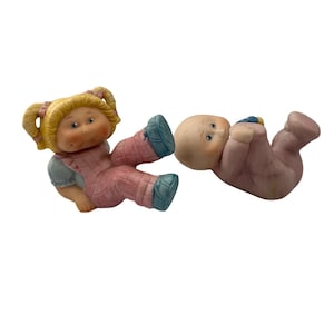 May include: Two vintage Cabbage Patch Kids figurines. One doll is wearing a pink jumpsuit and blue shoes, with blonde pigtails. The other is a baby in a pink onesie with a blue and yellow hat. Both are lying down.