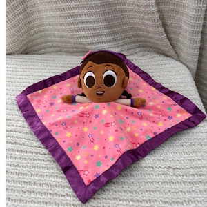 May include: A plush baby blanket featuring a cartoon character's head and arms. The pink blanket has a star pattern and a purple satin border. The character has brown skin, brown hair, and a pink bow.