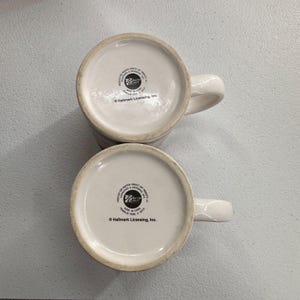 Puede incluir: Se muestran dos tazas de cer&aacute;mica blanca con asas curvas desde la parte inferior. La base de cada taza es blanca con un dise&ntilde;o circular y texto. Las tazas son probablemente para caf&eacute; o t&eacute;.