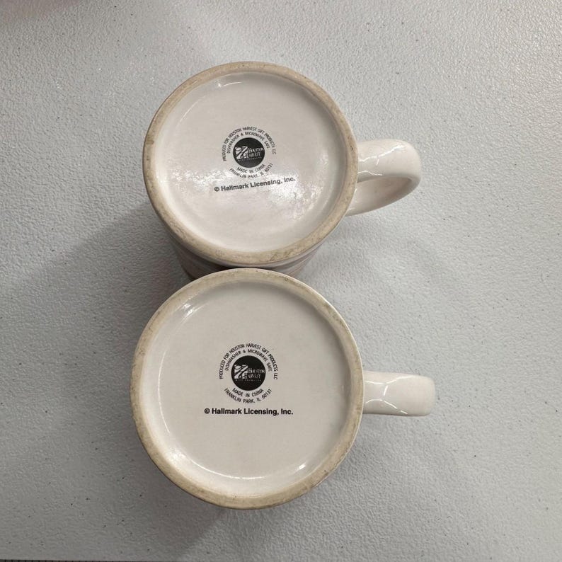 Puede incluir: Se muestran dos tazas de cer&aacute;mica blanca con asas curvas desde la parte inferior. La base de cada taza es blanca con un dise&ntilde;o circular y texto. Las tazas son probablemente para caf&eacute; o t&eacute;.