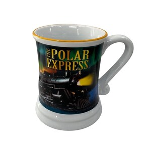 May include: A white ceramic mug featuring a graphic of a black steam locomotive and the text THE POLAR EXPRESS in gold lettering. The mug has a yellow rim, a curved handle, and a wide base, with a dark background depicting a night sky scene.
