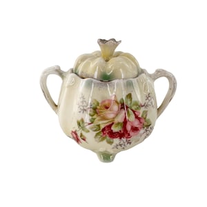Pode incluir: Açucareiro de cerâmica creme com tampa, com um design floral de rosas em tons de rosa e vermelho. O açucareiro tem duas alças e uma tampa decorativa com um pequeno botão. É um item vintage.