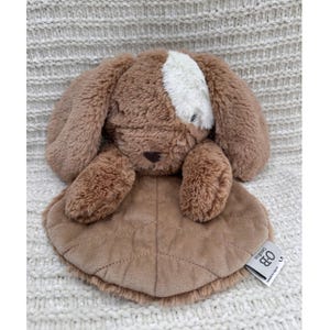 May include: A brown plush bunny toy with a white patch on its head and long floppy ears. The bunny has a soft, textured body and a quilted, leaf-shaped blanket. The toy is designed for babies and young children.