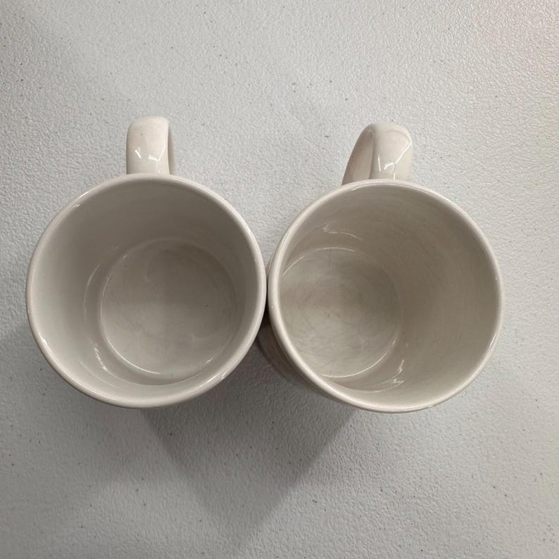 Puede incluir: Dos tazas de cer&aacute;mica blanquecina con asas curvas se colocan una al lado de la otra. Las tazas tienen un dise&ntilde;o sencillo y cl&aacute;sico, con un acabado liso y brillante. El interior de cada taza es de un tono ligeramente m&aacute;s oscuro de blanquecino.