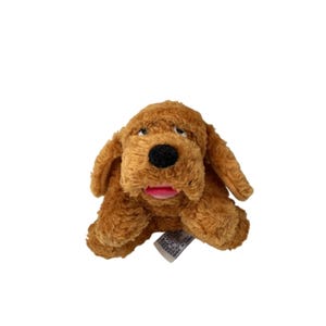 May include: A plush toy shaped like a brown dog with long floppy ears, a black nose, and an open mouth showing a pink tongue. The stuffed animal has soft, textured fur and a small white fabric tag attached to the bottom right side of its body.
