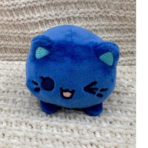 May include: A plush, blue cat-shaped toy with teal inner ears, black whiskers, and a pink mouth. The toy has small, rounded feet and is set against a textured, off-white background.