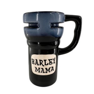 May include: A ceramic mug with a black base and a dark blue top section. The mug features a prominent black handle and a white rectangular plaque on the front with the black embossed text HARLEY MAMA. The design has a distinct tiered shape.