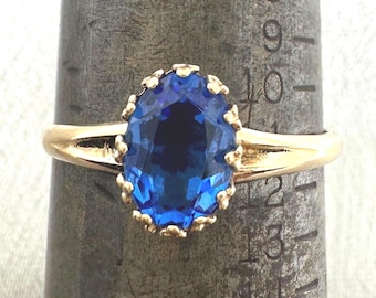 May include: A gold ring featuring an oval-cut blue gemstone set in a scalloped prong setting. The ring is positioned on a metal ring mandrel, with the band resting between the size 10 and 11 markings on the graduated scale of the tool.