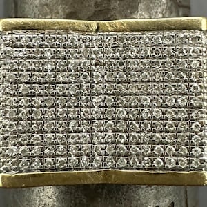 Puede incluir: Un anillo rectangular con una banda dorada y una superficie cubierta de pequeñas piedras brillantes. El diseño del anillo presenta un borde dorado que enmarca la matriz de piedras, creando un accesorio lujoso y llamativo.