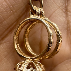 May include: A gold-coloured pendant featuring two intertwined rings, connected by a decorative bail. The rings have a textured design, and the pendant has a floral-shaped base. The jewellery is likely a charm or a small necklace pendant.