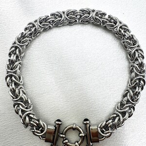May include: A silver-toned chain necklace featuring a complex Byzantine weave pattern. The necklace is secured with a circular toggle clasp mechanism that connects two cylindrical end caps, creating a continuous circular loop design.