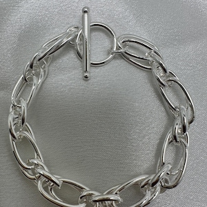 May include: A silver-colored chain bracelet with an oval link design. The bracelet features a toggle clasp closure with a circular ring and a straight bar. The jewelry is displayed on a light gray, satin-like surface.