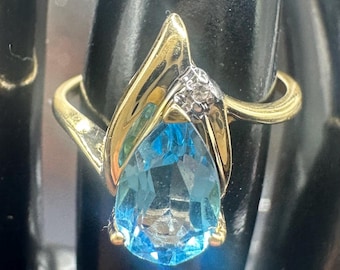 May include: A gold ring with a pear-shaped, light blue gemstone. The ring features a gold band and accents, with a small cluster of diamonds near the top of the gemstone. The ring is set against a dark background.