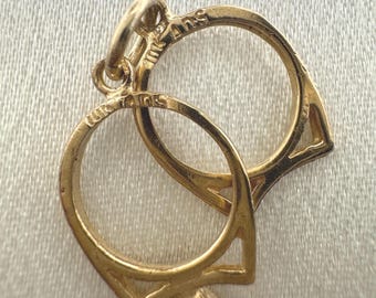 May include: A gold pendant featuring two interlocking circular frames, each with a pointed base. The metal surface is engraved with the text 10K ANS. The pendant includes a small circular jump ring at the top for attachment to a chain or cord.