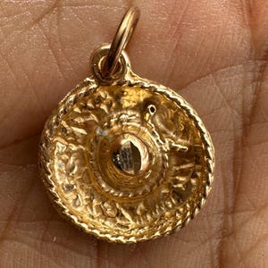 Puede incluir: Un colgante dorado con un diseño circular. El colgante tiene una superficie texturizada con detalles intrincados y un pequeño lazo en la parte superior para sujetarlo a una cadena. El centro del colgante tiene una pequeña piedra de color oscuro.