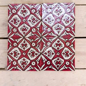 Peut inclure: Quatre carreaux de céramique carrés avec un motif floral rouge et blanc répétitif. Les carreaux sont disposés ensemble, mettant en valeur le motif. Le fond est une surface en bois de couleur claire.