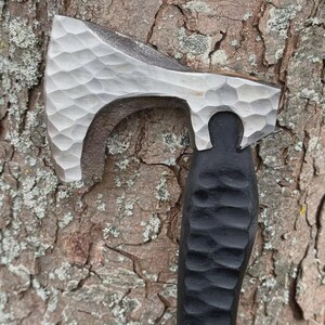 May include: A hand-forged axe with a textured, silver-colored head and a black, carved wooden handle. The axe head has a unique, dimpled pattern. The handle is carved with a repeating, curved design. The axe is set against a textured, brown tree trunk.