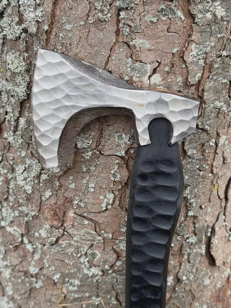 May include: A hand-forged axe with a textured, silver-colored head and a black, carved wooden handle. The axe head has a unique, dimpled pattern. The handle is carved with a repeating, curved design. The axe is set against a textured, brown tree trunk.
