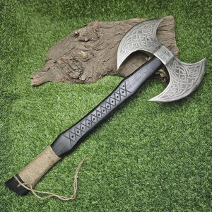 May include: A double-bladed axe with a silver head and a black handle. The axe head features intricate Celtic knot designs. The handle is decorated with diamond-shaped patterns and wrapped with twine near the base. The axe is displayed on a green surface.