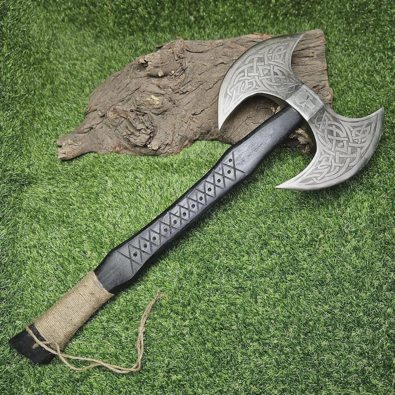 May include: A double-bladed axe with a silver head and a black handle. The axe head features intricate Celtic knot designs. The handle is decorated with diamond-shaped patterns and wrapped with twine near the base. The axe is displayed on a green surface.